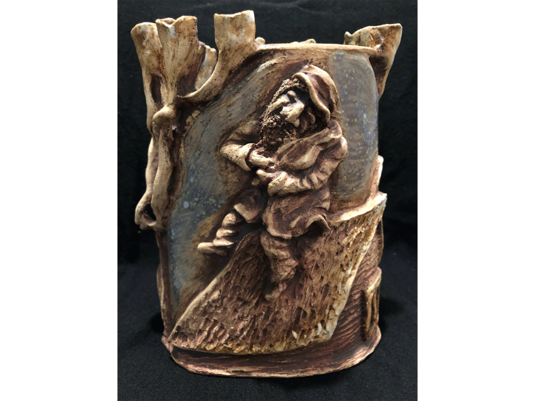 The artwork titled "Why Do We Stay Up There If It's So Dangerous?" was created in 2019 by Julia Ilyutovich. The image is a small ceramic vase with earthy light blue and brown colors. A relief sculpture of a bearded man in a large coat and hat is centered on the front of the vase. The man is a fiddler, holding his violin up to his chin. The ceramic artwork is 8 inches tall, 3 inches wide, and 2 inches deep.
