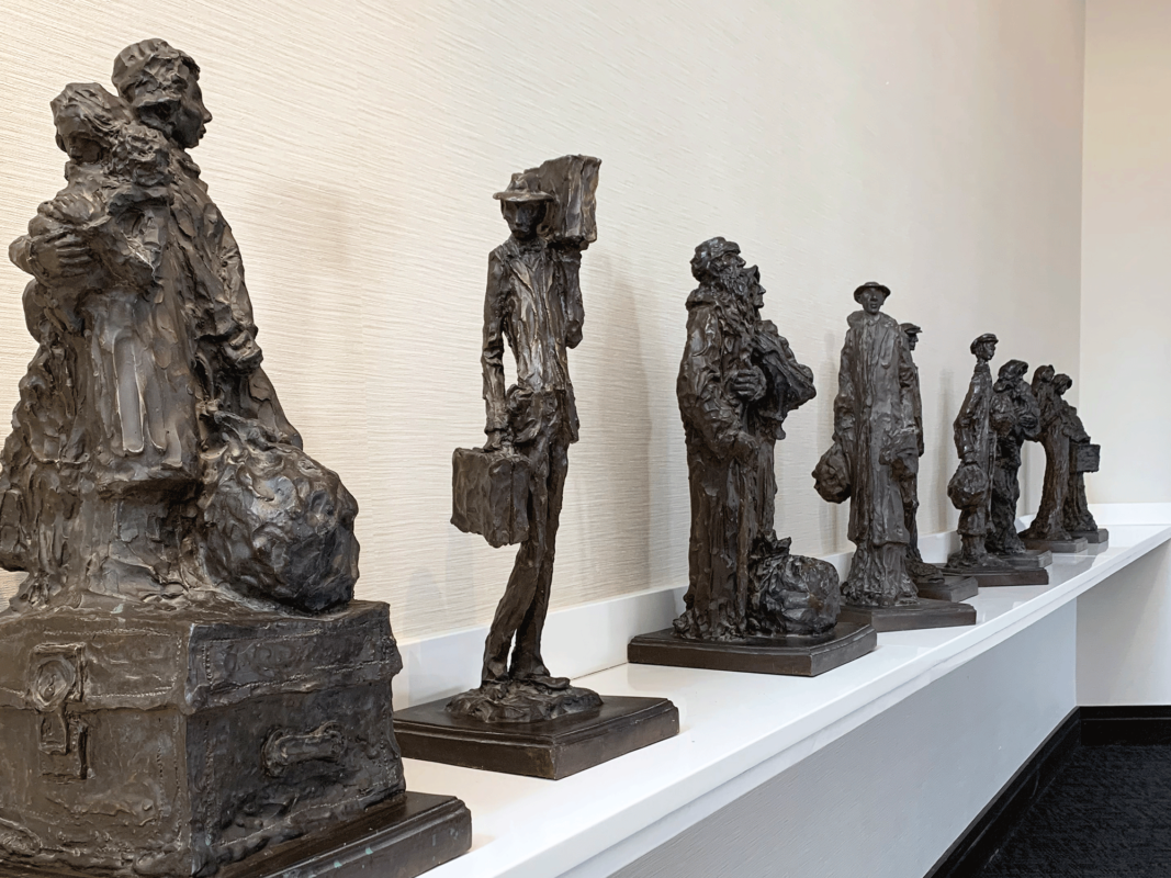 The sculpture installation titled "Immigrant at Ellis Island" was created in the early 1970s by Phillip Ratner. The image is a digital photograph of nine maquettes on display on a bright white shelf in a well-lit museum space. The maquettes are made of a dark clay or plaster material, each one depicting a 20th century European immigrant at Ellis Island. The sculptures are slightly abstract and extremely textured.
