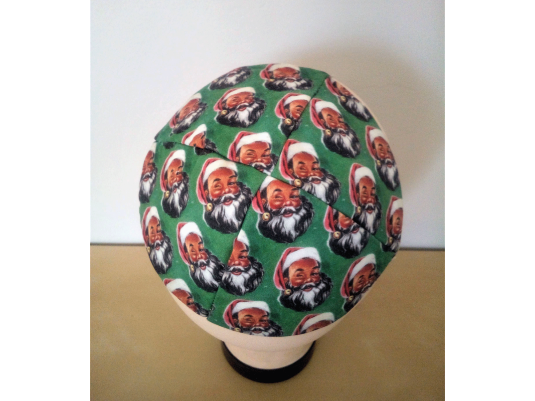 The artwork titled "Santa Yarmulke" was created in 2018 by Jacob Rath. A brimless cap called a kippah is overrun with repeating images of Santa Claus's face on a green background. The fabric kippah measures 5.5 inches in diameter.