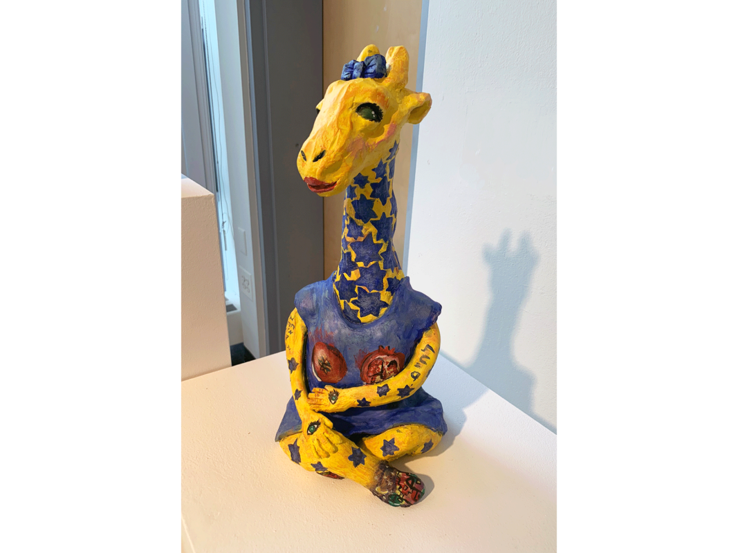 The artwork titled "Jewraffe" was created in 2019 by Cassandra Clark. The ceramic artwork is 14 inches tall, 12 inches wide, and 16 inches deep. A sculpture of a anthropomorphic female giraffe sits cross-legged on a white shelf in a well-lit museum space. Instead of black spots, the giraffe has many blue stars covering her neck. There are also well-spaced blue stars and Hebrew letters on the giraffe's arms and legs, and she is wearing a blue short sleeved dress with pomegranates painted over her breasts. A small multi-colored cityscape creates the illusion of shoes on her feet. A blue bow sits between the giraffe's ears, and she is sporting bright red lipstick.