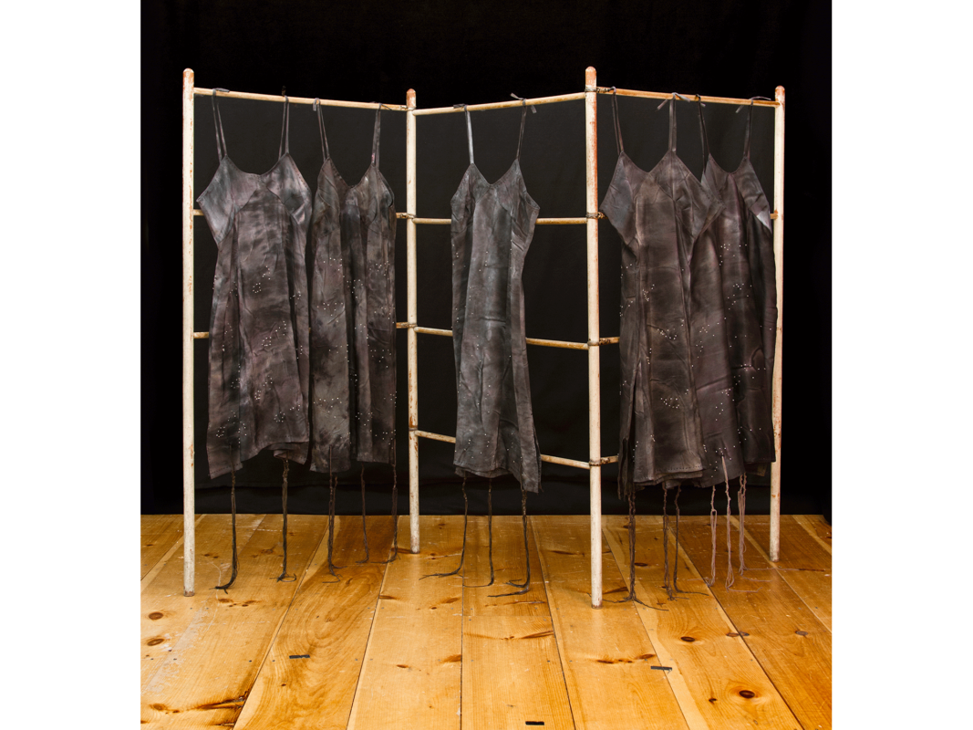 The artwork titled "Tallit Katan" was created in 2013 by Rachel Kanter. Five dark gray silk dresses hang from an old wooden drying rack. The artwork uses hand dyed silk with glass beads and a vintage drying rack. The piece is 65 inches long, 48 inches wide, and 48 inches deep.