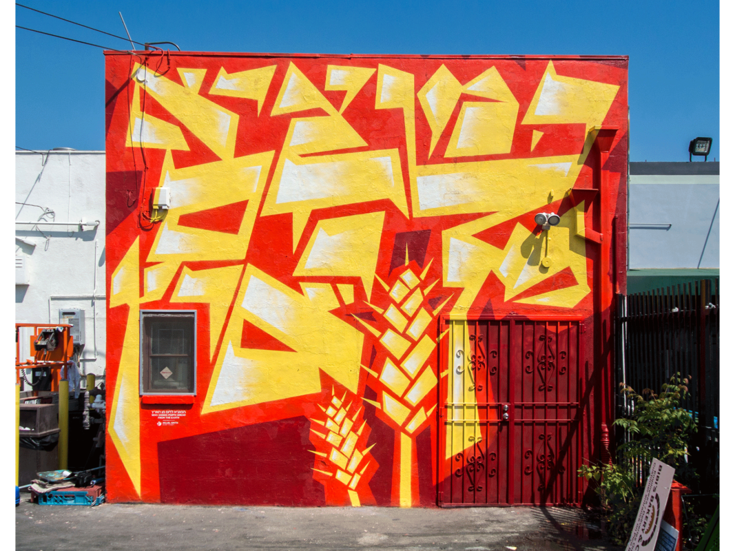 The artwork titled “Hamotzi Lechem Min Ha-aretz" was created in 2016 by Hillel Smith. The image is a photograph of painted mural, 14 inches by 11 inches. The back wall of a two-story kosher bakery is painted with vivid red, orange, and yellow geometric words “HaMotzee Lehem Min ha’Aret," meaning, "who brings forth bread from the earth." These enormous words are placed next to two stylized stalks of wheat, of bright, white-highlighted yellow against a deep orange and shadows of dark reddish-violet.