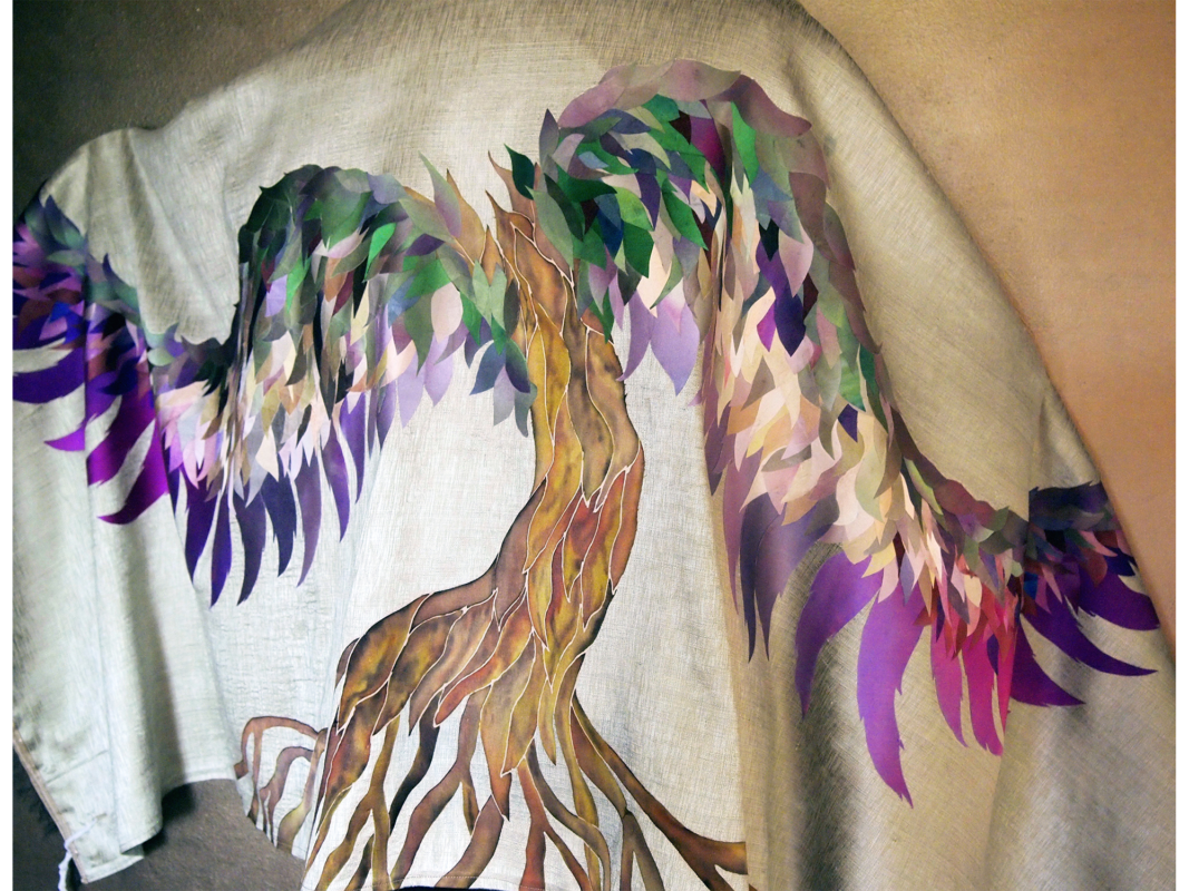 The artwork titled "Wings of Peace Tallit" was created in 2019 by Diane Fredgant. The image is an off-white tallit embroidered with a large tree with green, biege, and purple leaves sprouting from its branches. The branches and leaves create the illusion of wings. The artwork is hand-painted silk and fabric, 24 inches long and 72 inches wide.