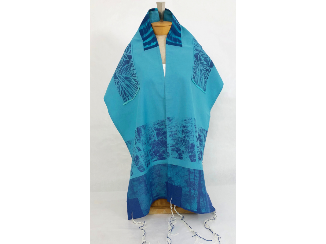 The artwork titled "Trees Tallit" was created in 2019 by Ruth Simon McCrae. The image is a turquoise blue shawl with blue threads interwoven in the white fringes. The artwork is a photo silkscreen and stencil on cotton and linen, 15 inches by 78 inches.